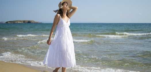 beautiful young woman walking on sandy beach wearing a hat and a white dress enjoying the sun